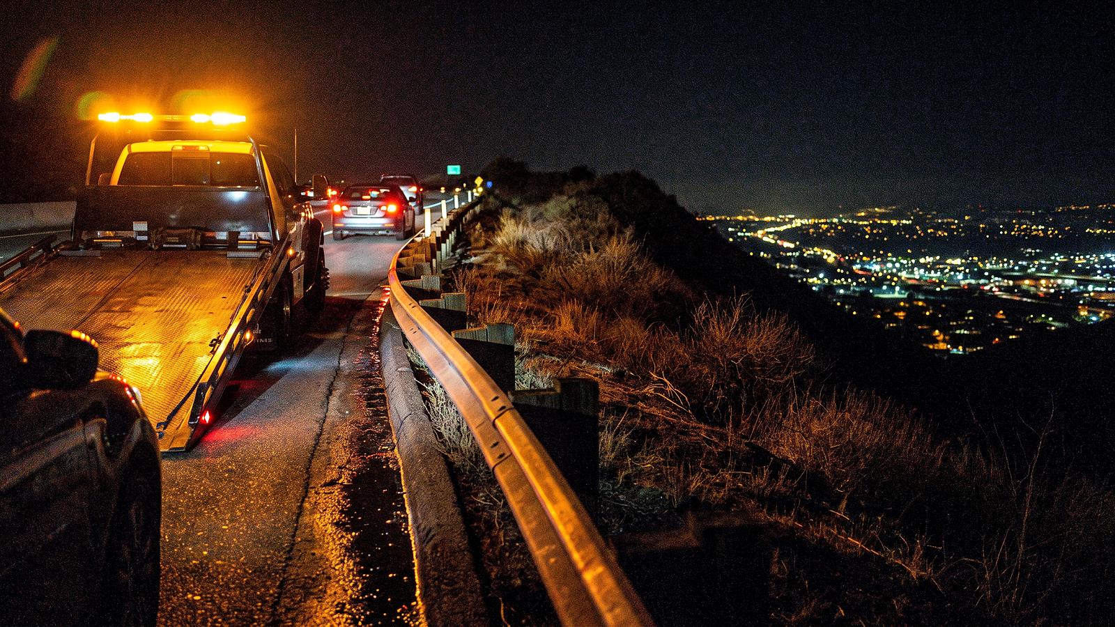 A tow truck's amber light bar on a freeway shoulder at night above a valley of lights