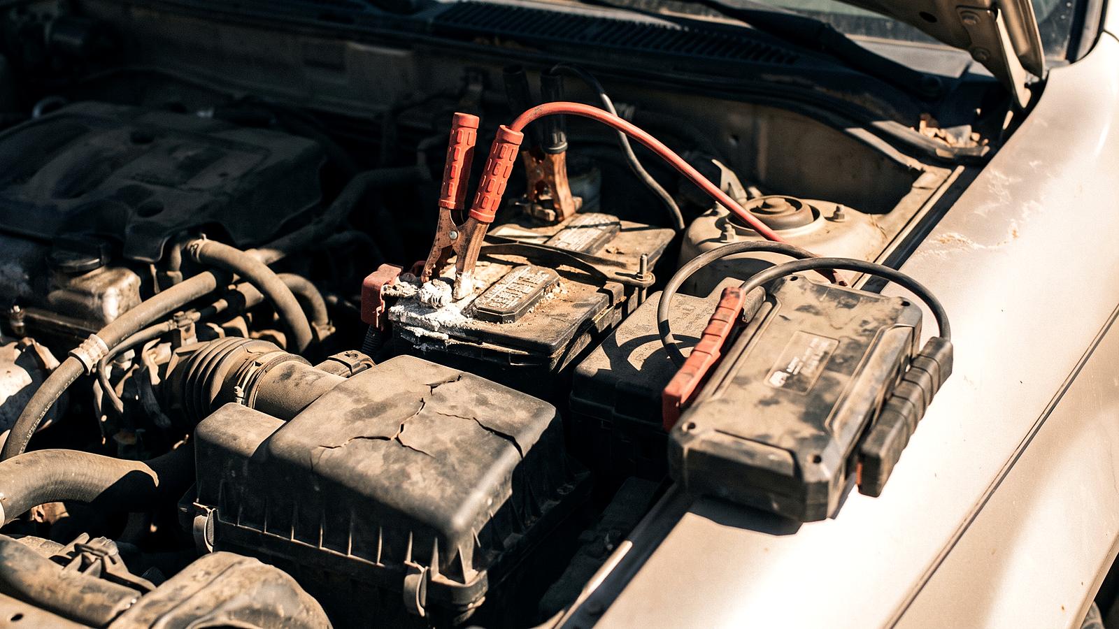 Jumper cable clamps on a car battery terminal under an open hood in hard afternoon sun